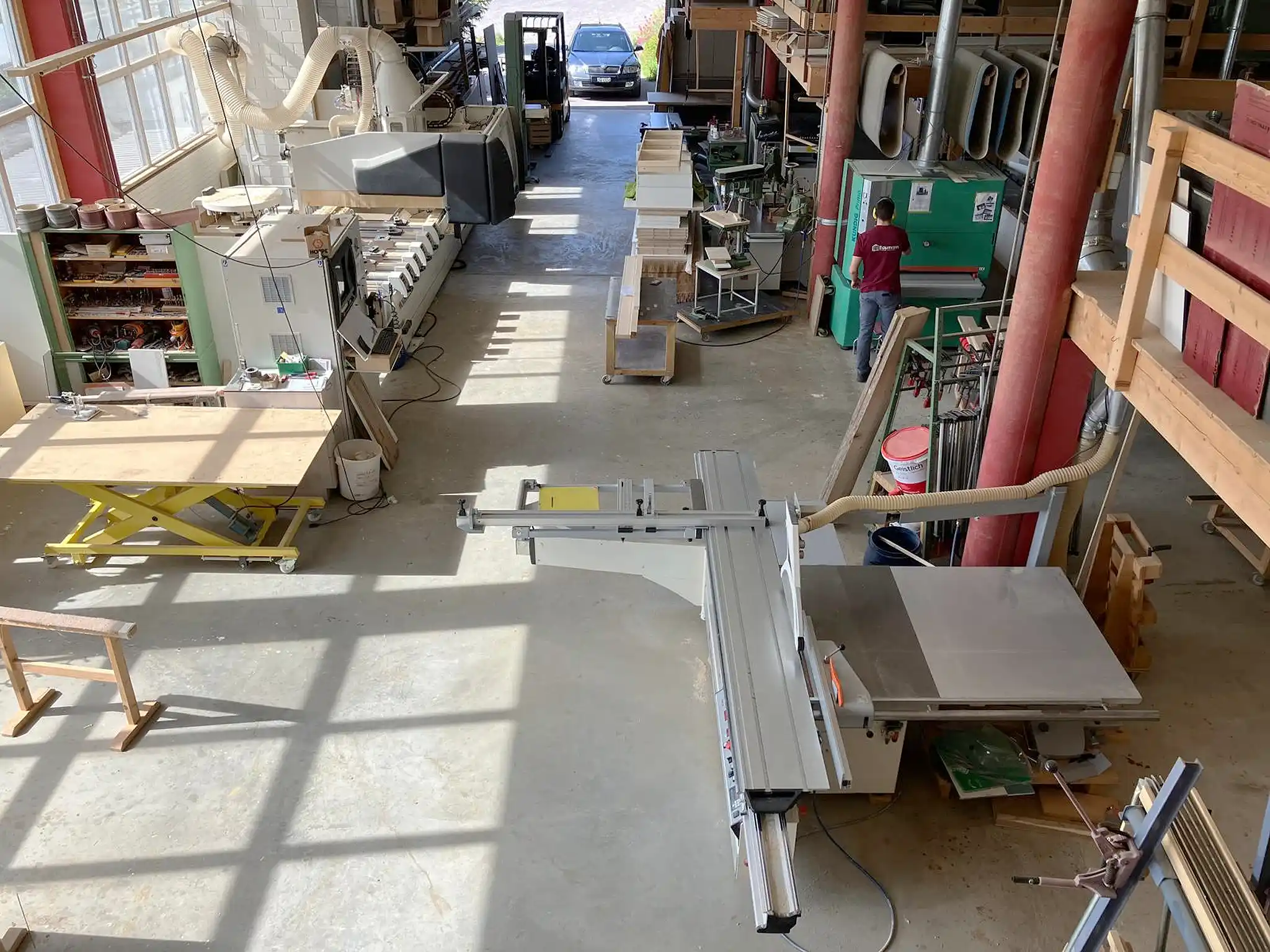 Blick von oben in die Holzwerkstatt mit CNC-Anlage, Formatkreissäge, Breitbandschleifmaschine und zentraler Absaugtechnik für die professionelle Holzverarbeitung. Blick von oben in die Holzwerkstatt mit CNC-Anlage, Formatkreissäge, Breitbandschleifmaschine und zentraler Absaugtechnik für die professionelle Holzverarbeitung.